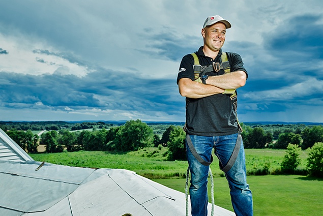 Proud GAF Energy Employee standing on roof with blue sky and rolling green behind